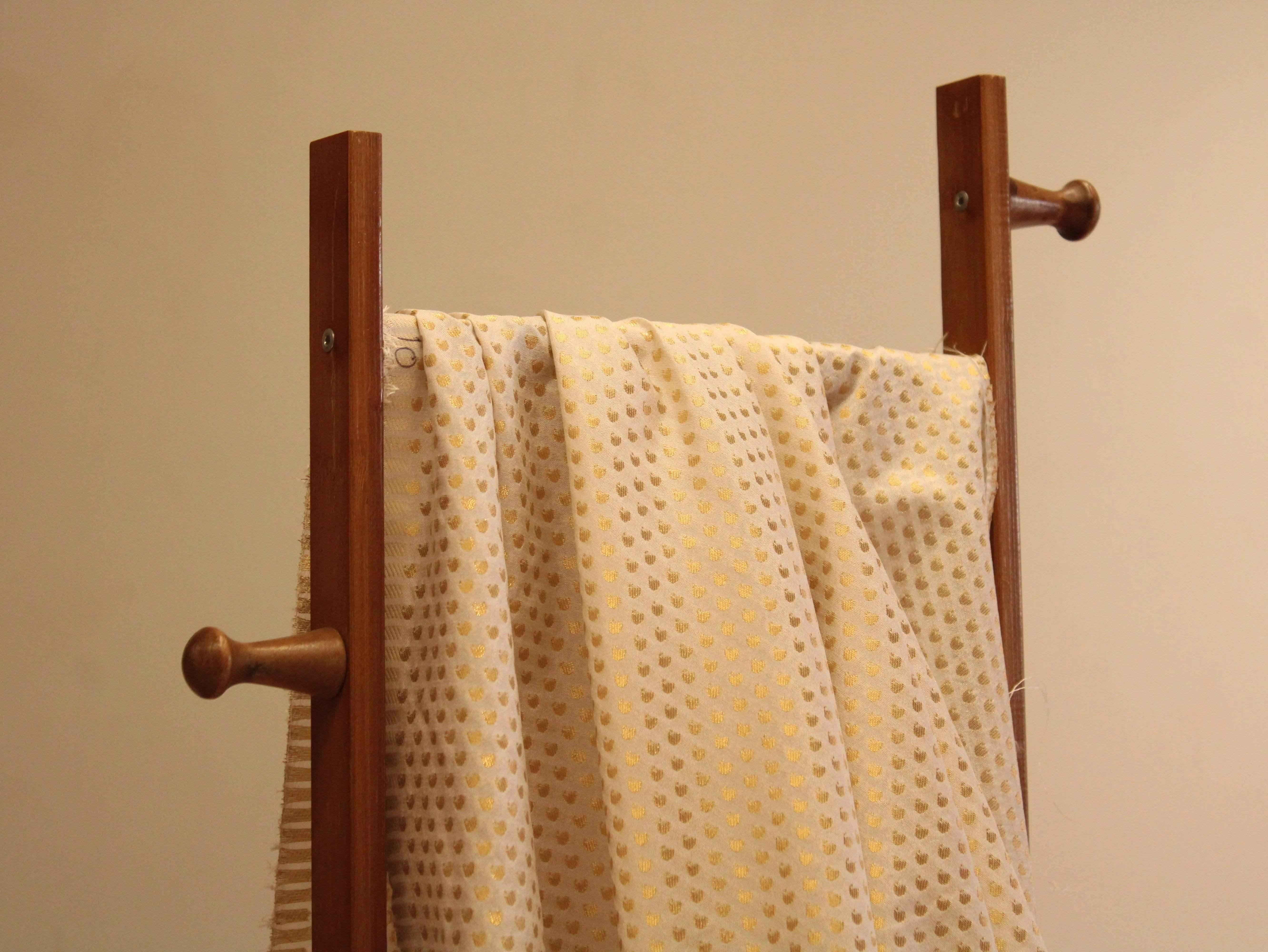 Off-white Banarasi brocade fabric with golden motifs draped on a wooden stand