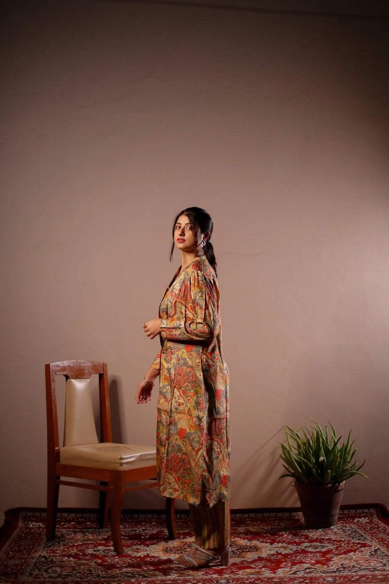 Woman in a floral printed co-ord set with long blazer and pants, standing indoors on a patterned rug next to a wooden chair and potted plant.