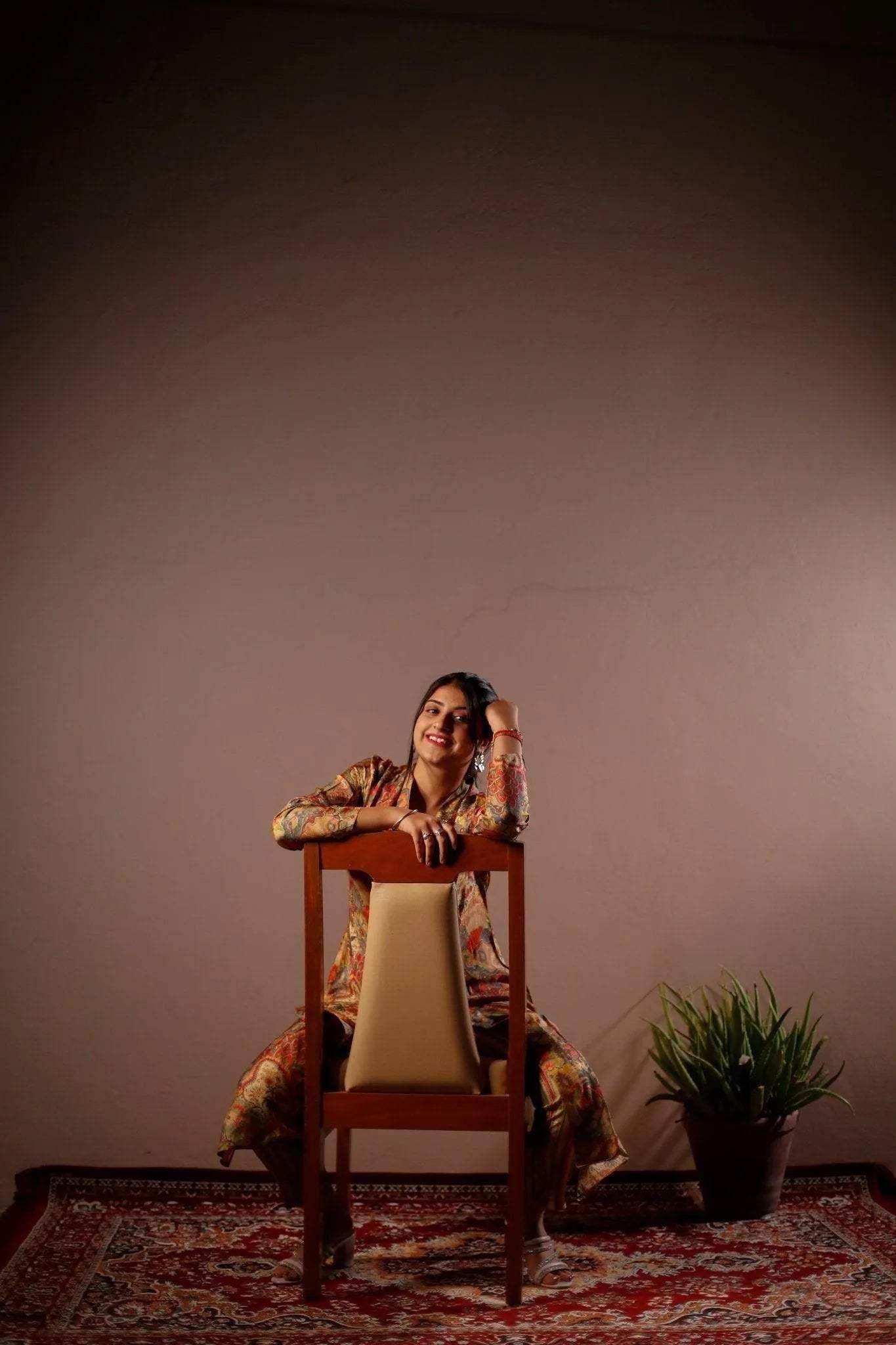 Woman in floral blazer and pant co-ords set sitting on chair, indoor with a patterned rug and plant