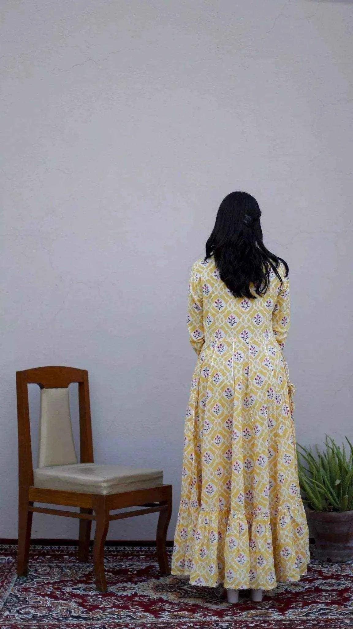 Woman in a yellow mulmul cotton floral gown standing indoors near a chair and potted plant